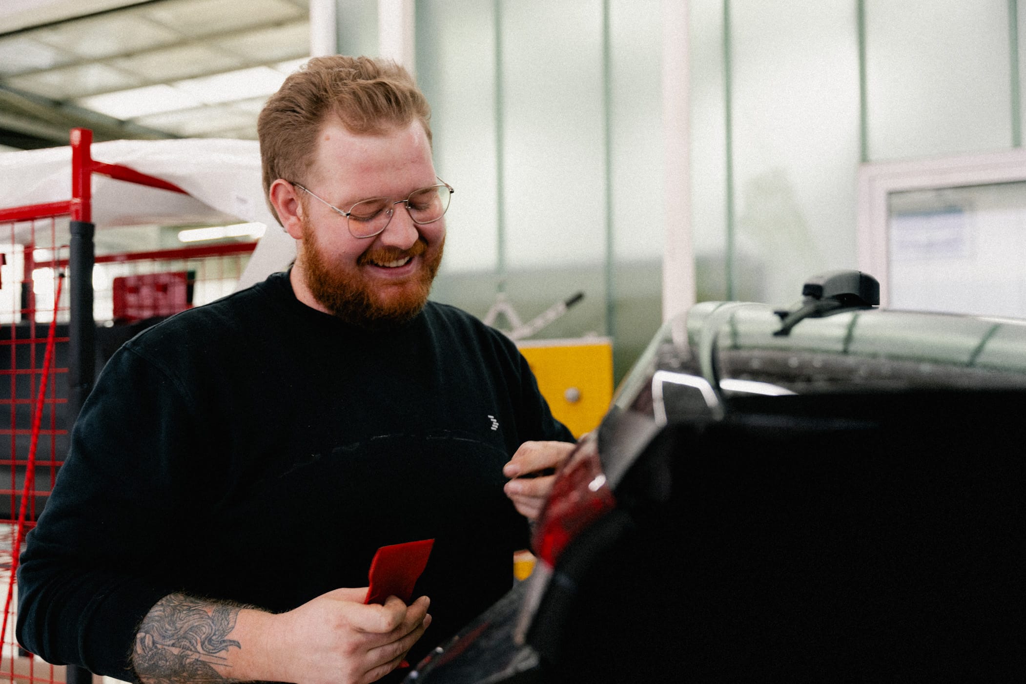 Eine Portraitaufnahme eines Mitarbeiters des Autohauses in der Werkstatt. Er steht vor der Heckklappe eines Autos und führt Reparaturen durch. In seiner rechten Hand hält er ein Werkzeug. Außerdem lächelt er und ist bekleidet mit einem schwarzen AVEMO Pullover.